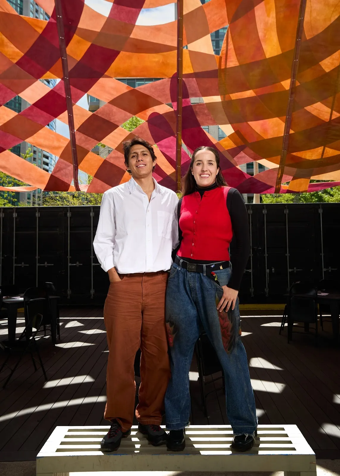 Two people pose together, smiling in front of a canopy-like piece outdoors. The canopy is made from strips of red and orange fabric crossing over each other. Spots of sun illuminate the ground where the canopy has grid-like gaps.