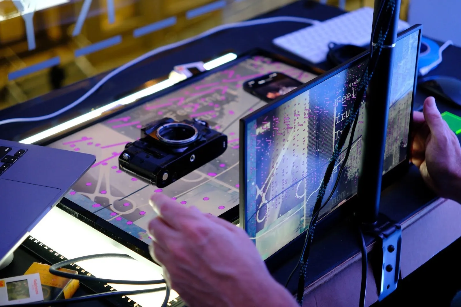 A close-up of a tabletop workspace with a disassembled camera body resting on a lightbox, surrounded by cables, a laptop edge, and a small monitor displaying glitchy graphics. Hands adjust equipment in the foreground.