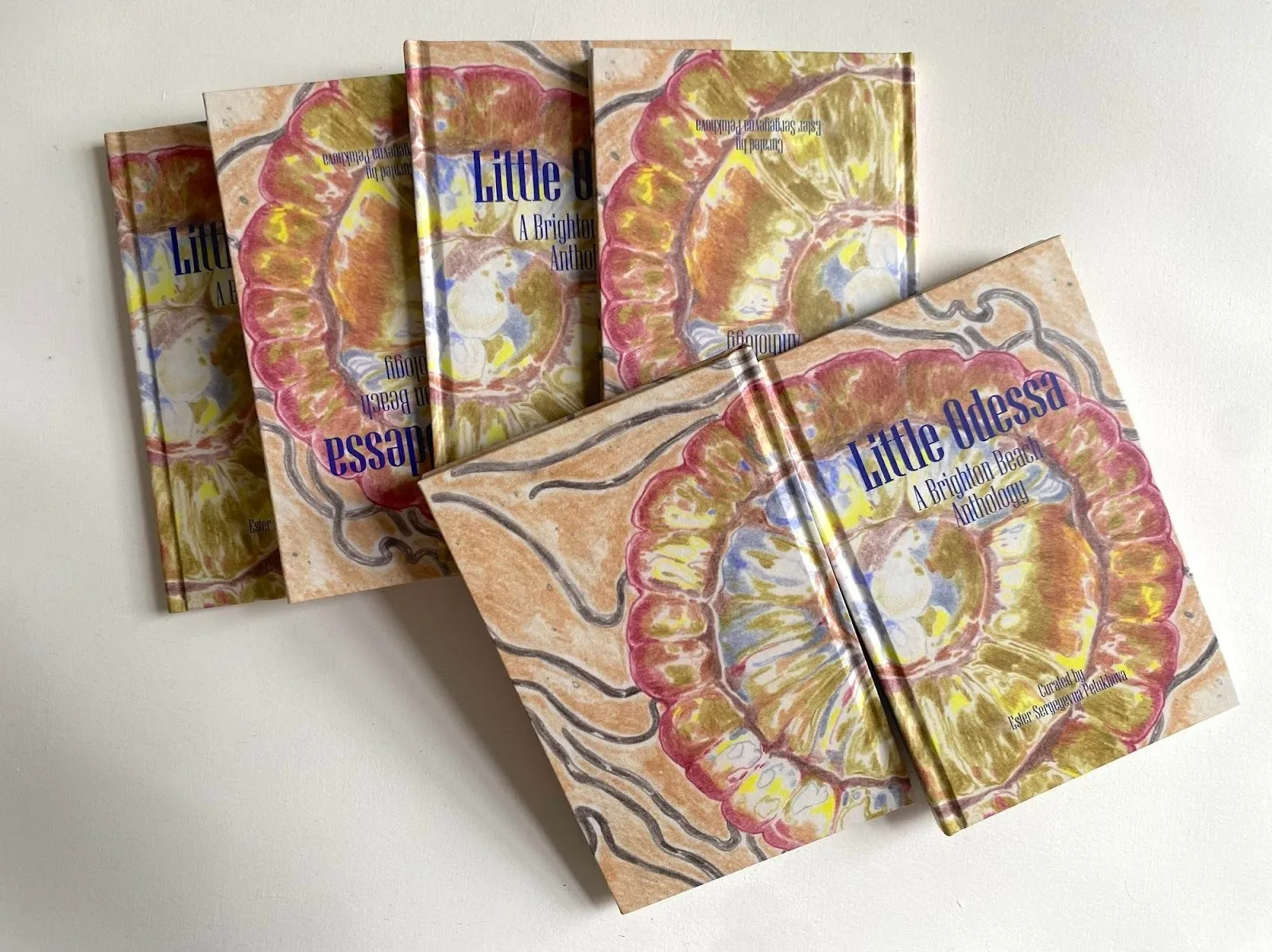 Photo of multiple copies of a book strewn about, stacked on top of each other. The cover art features a painting/drawing of an abstracted, iridescent flower. The book title reads "Little Odessa, A Brighton Beach Anthology" in blue sarif.