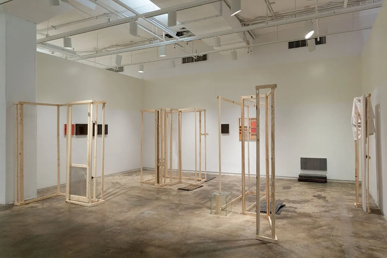 Small paintings line gallery walls. Four sparse, rectangular wooden structures are placed about the space, resembling crude, structural, wall-like frames.