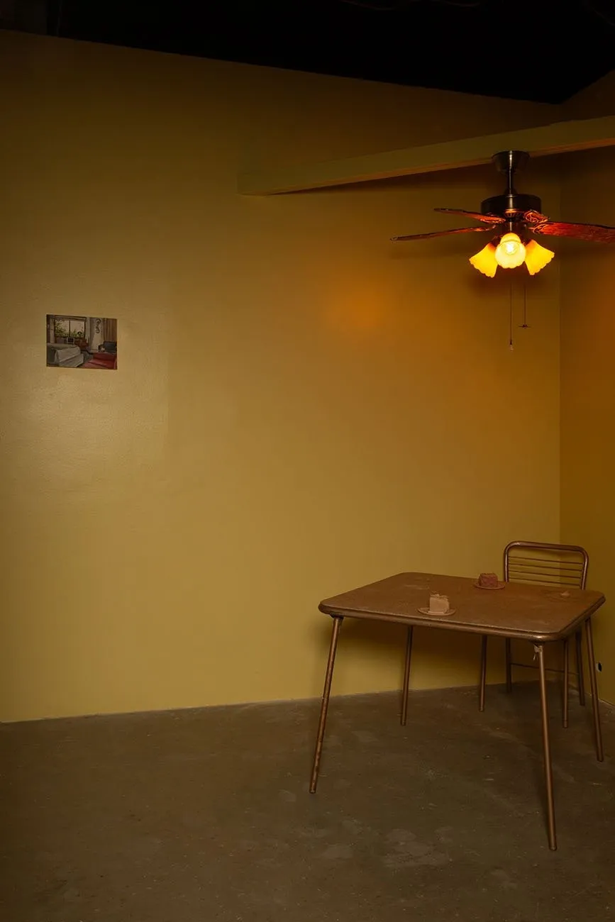 A yellow-painted room is dimly lit. A fan with lights hangs, illuminating warm light. A single, small image is pasted to the top left wall. A simple, small wood and metal table and chair rest in the corner. On the table, there are two slices of cake.