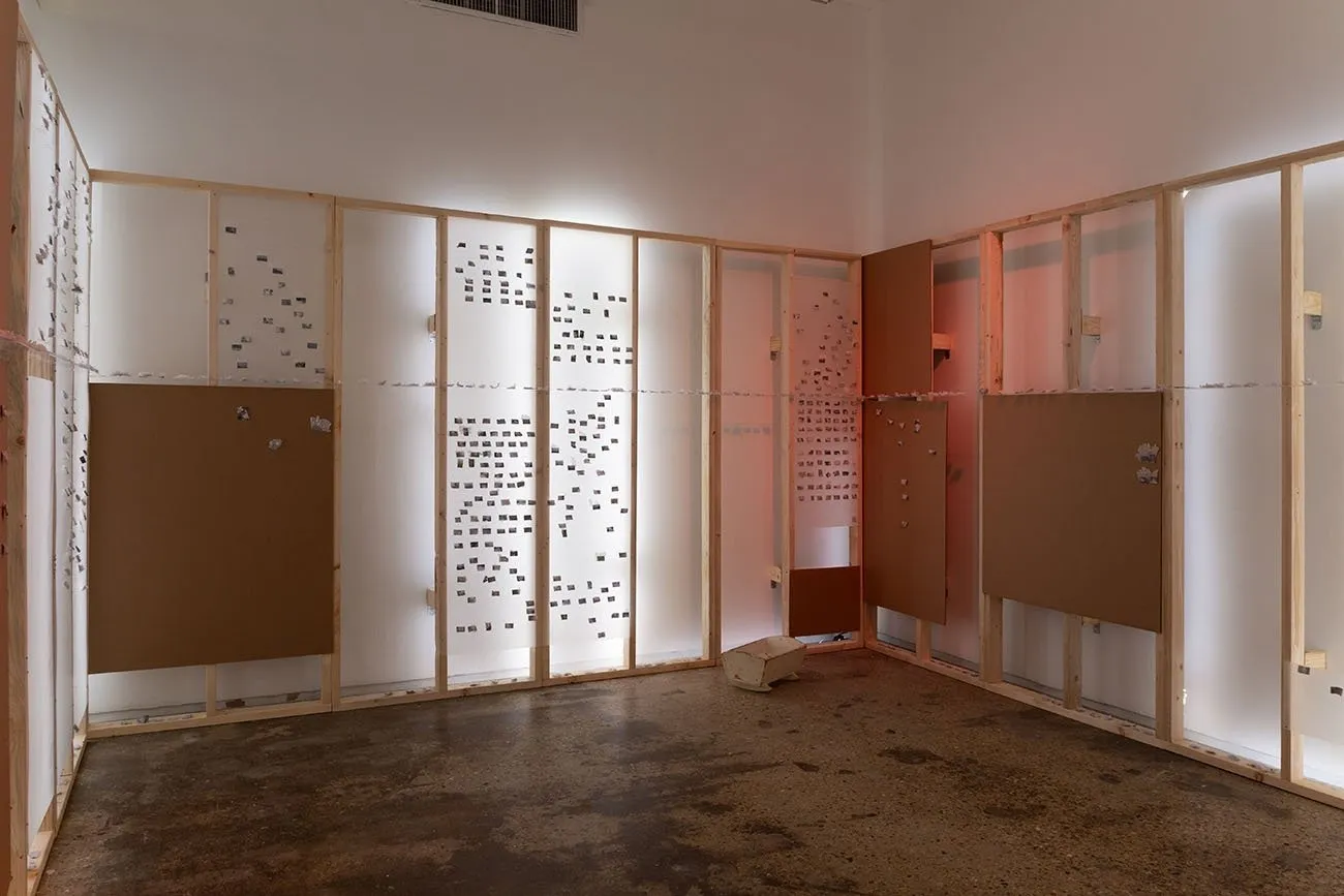 A white gallery room is lines with continuous wooden frames. Some frames have back lighting, producing a soft, white glow. Each frame has small media suspended within or wooden boards attached.