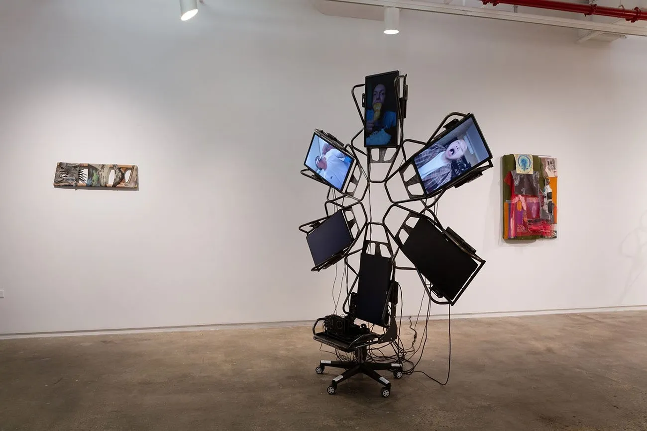 Two 2D works are mounted to a gallery wall. On the floor, a large sculpture made of monitors configured in an asterisk shape stand on a rolling chair with wires hanging about. Each monitor is either black or features a figure.