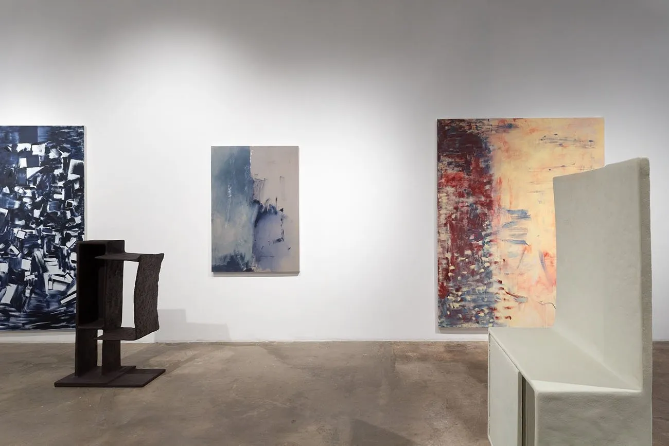 Paintings and sculpture populate a gallery room. Both painting and sculpture share geometric, square qualities. The paintings appear abstract and cubist, while the sculptures make simple, hollow, square forms.