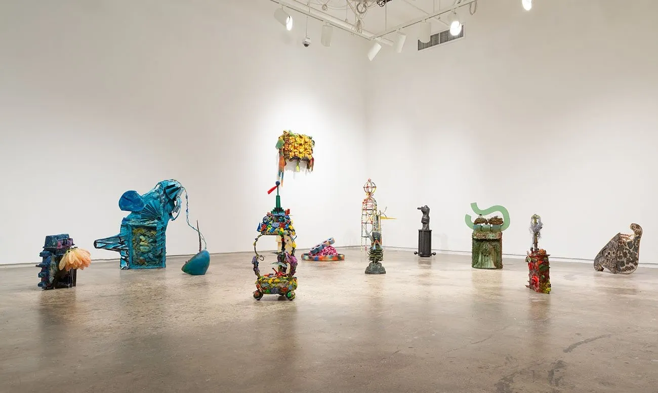Multiple small, colorful sculptures dot the floor of a gallery space. The sculptures appear as amalgamated parts with abstract, distended forms.