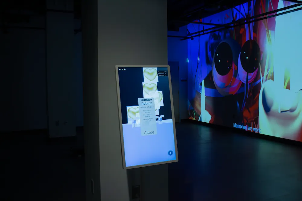 A standing touchscreen displaying a “Donate Robux!” pop-up menu in a dim gallery, with a large projection of a wide-eyed character playing behind it.