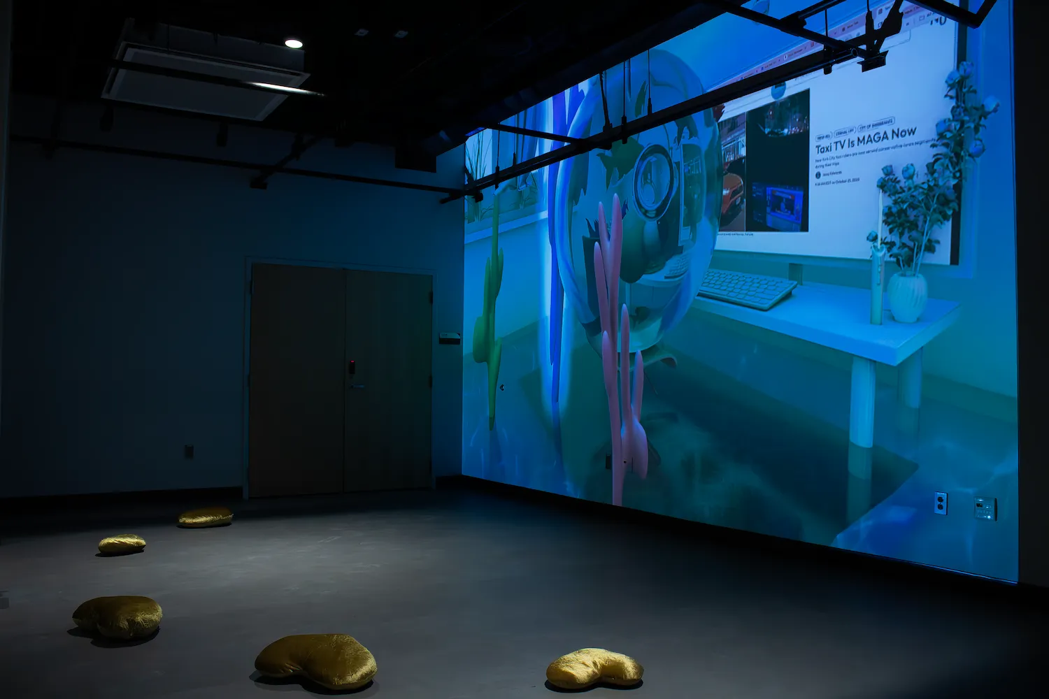 A dark gallery room with a large blue-toned digital projection showing a surreal desktop scene; several gold heart-shaped floor cushions are spread across the floor.