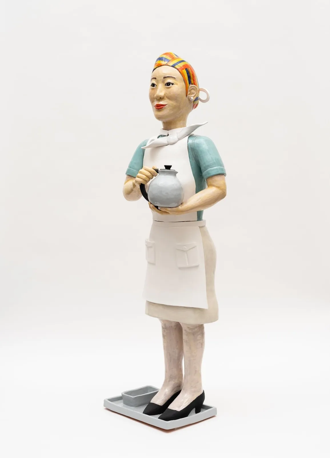 A ceramic sculpture of a woman in a flight attendant's blue and white outfit. She holds a metal teapot in both her hands. She smiles and looks outward. Her hair is made of red, yellow, and blue stripes.