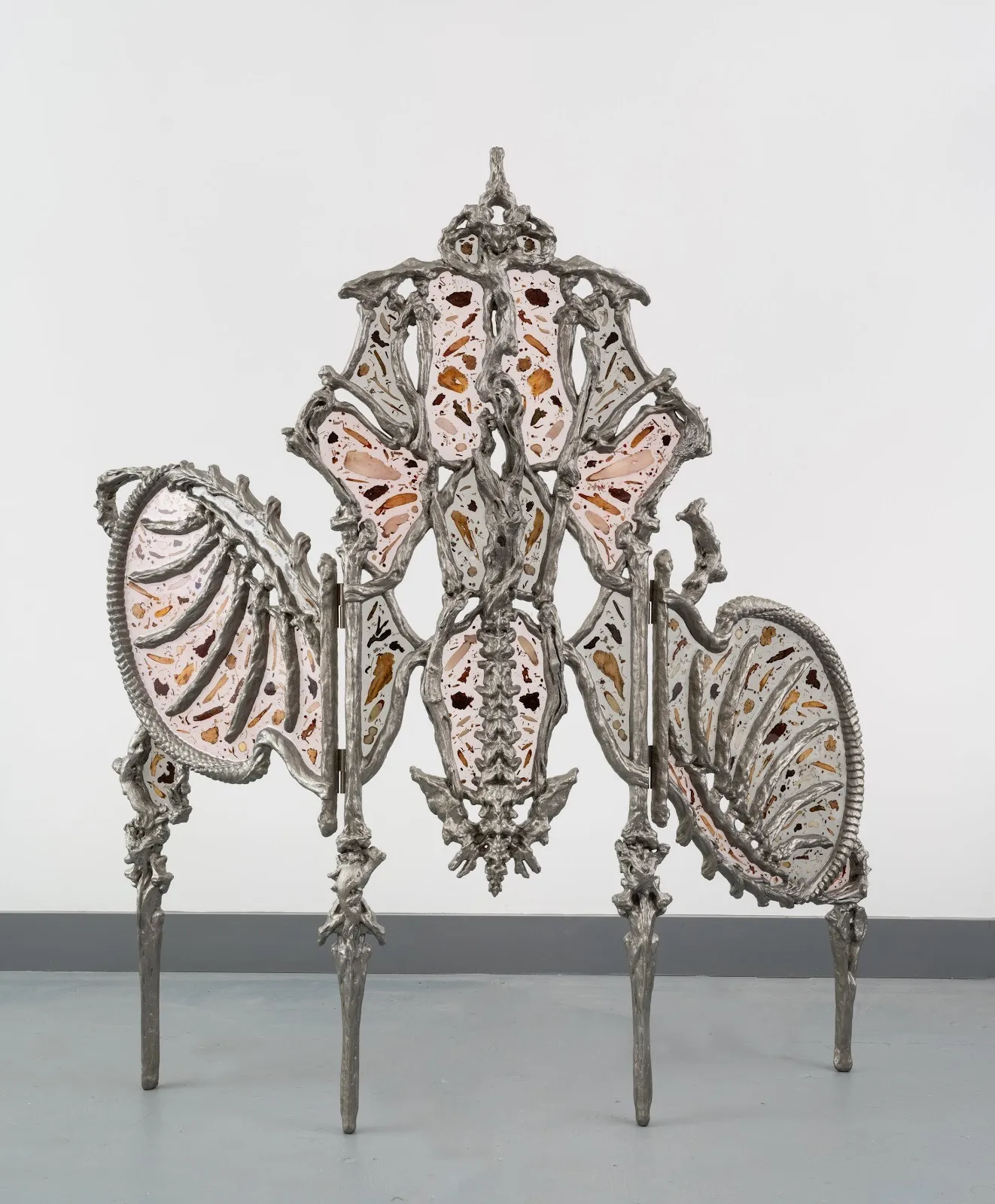 An organic yet alien sculpture rests on four bony legs. An intricate frame of chromelike, reptilian, bone structures support translucent, fossil-like panes stretched between. The piece is vaguely symmetrical with a central spinal column-like middle.