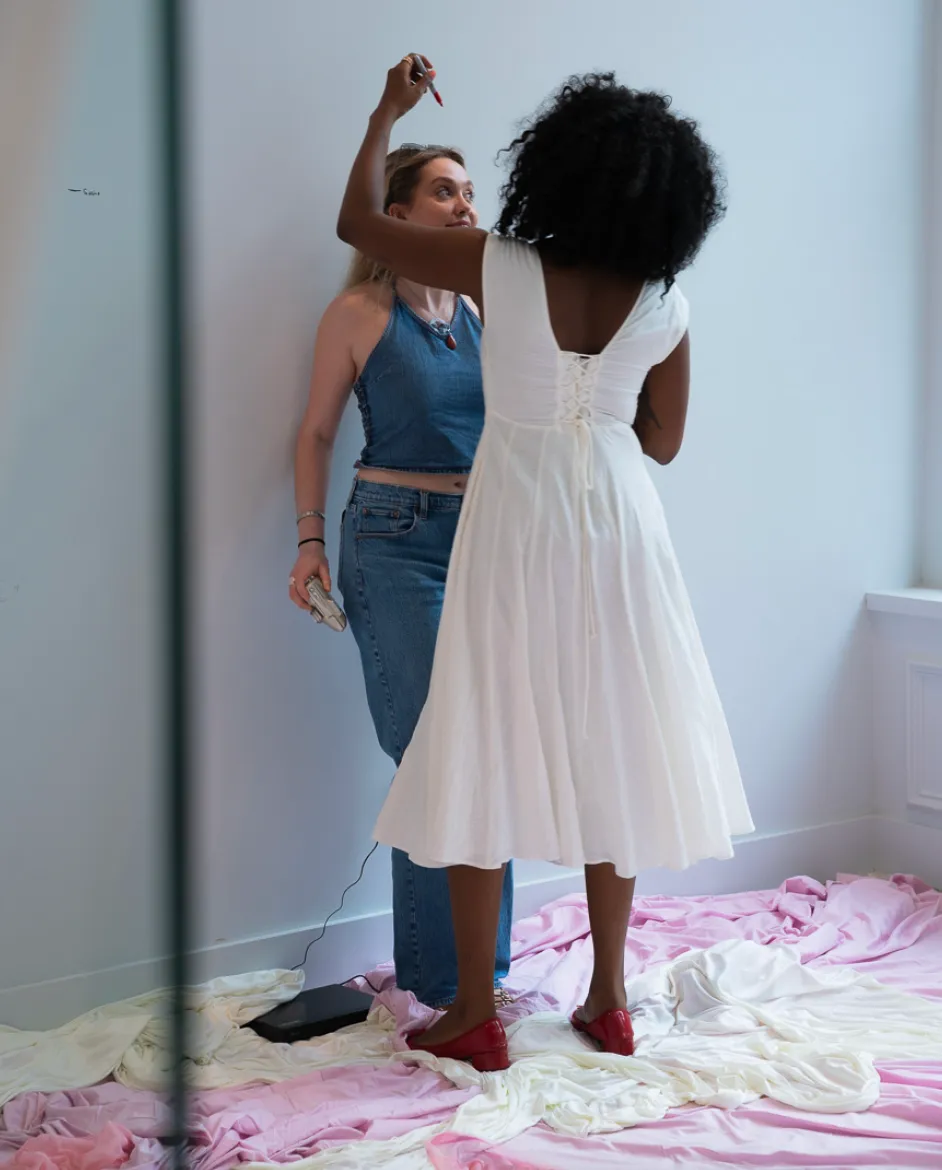 A person measures the height of another person with a red sharpie against a white wall. They stand on scattered, piled pink and white fabrics on the floor.