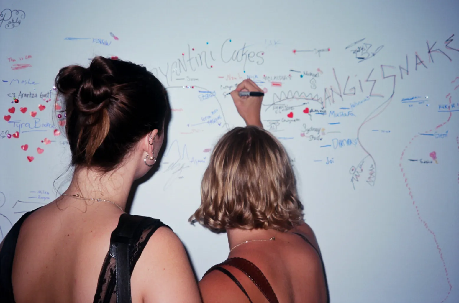 Two people face a wall and write their signatures on it with a sharpie. The wall is filled with other people's signatures, each in different styles and sizes, some decorated with different colors, sparkles, and heart-shaped stickers.
