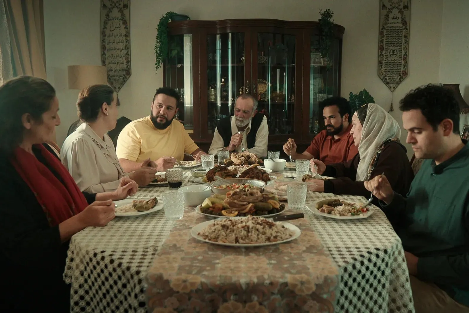 Seven people gather around a dining table in a dining room, eating multiple dishes of food and conversing with each other.