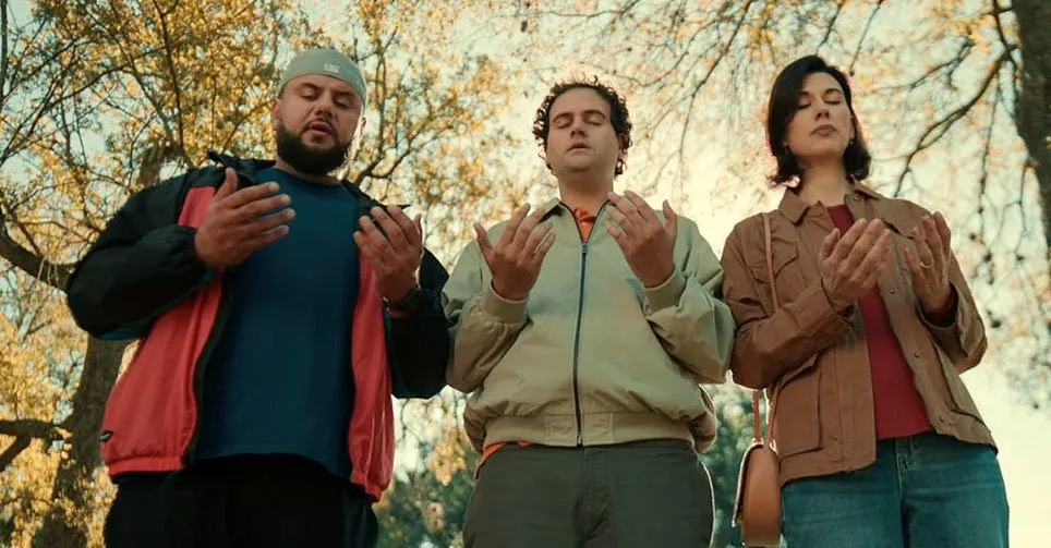 In a cemetery with many autumn trees, three people stand together and lift their hands, speaking prayers.