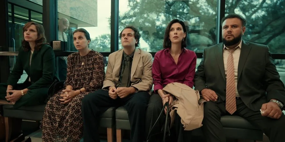 Five people sit in a line, their backs against a large window. They seem to be looking at something to the left of the camera, with some of them in slight distress.
