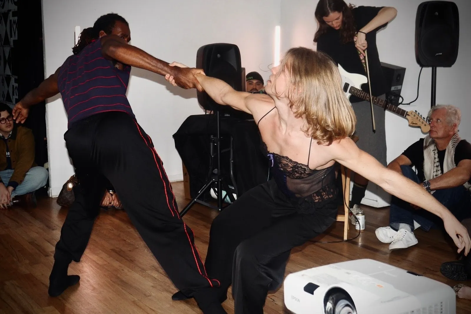 Two performers in motion holding hands mid-dance, with a musician playing guitar behind them and attendees seated on the floor.