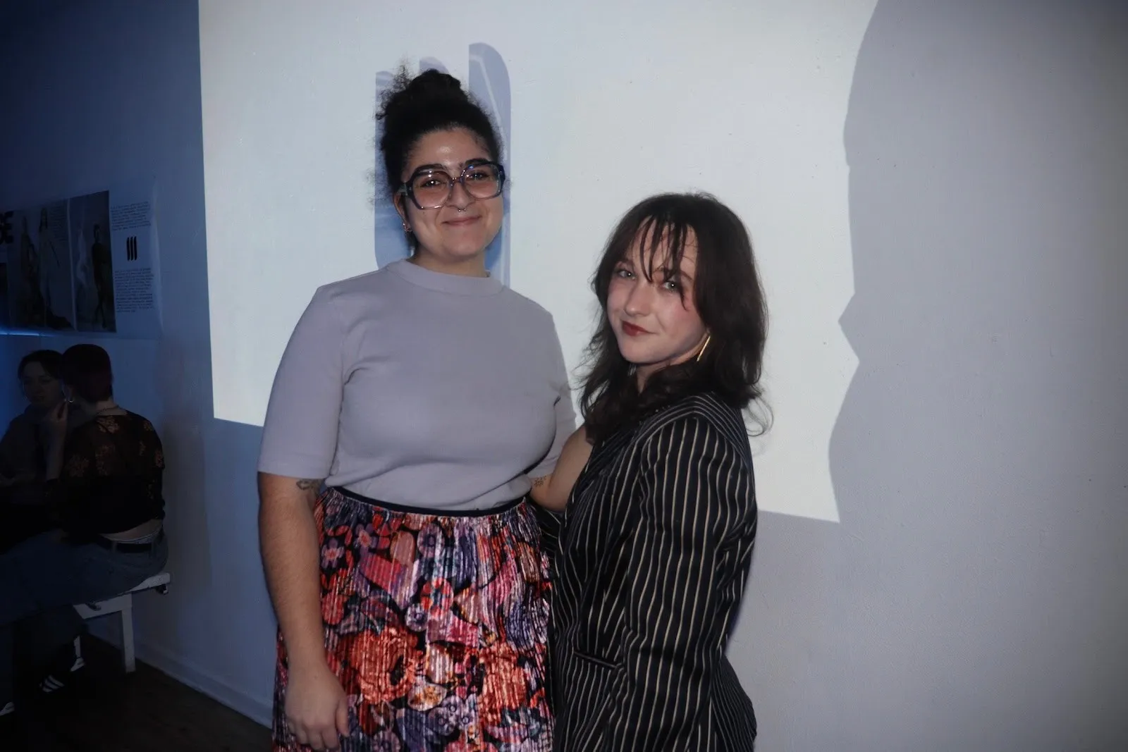 Two people posing together against a wall with projected light, one wearing glasses and a floral skirt, the other in a pinstripe jacket.