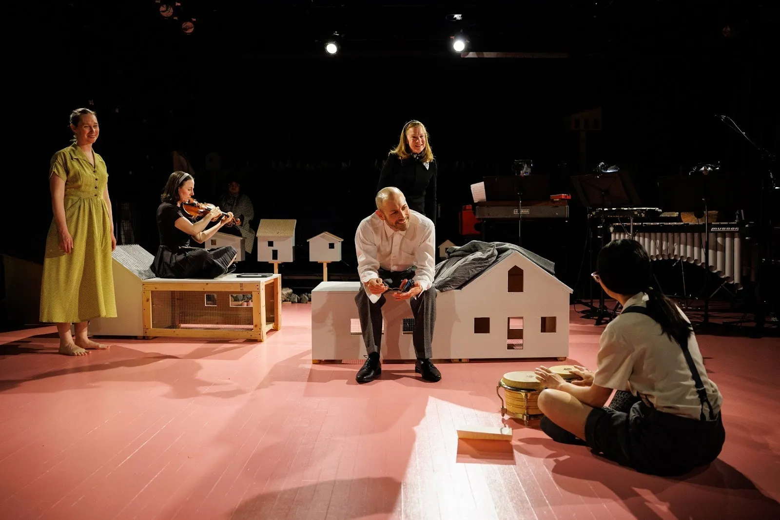 Five performers play various instruments and sing on stage. Props on the stage include benches and couches shaped like small houses. Instruments can be seen in the background.