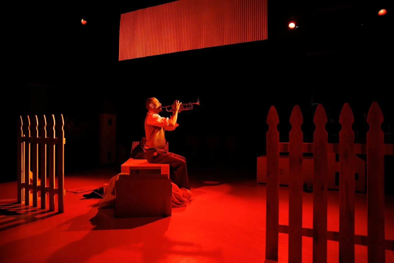 Under red lighting, a lone performer on stage sits on a prop bench and plays the trumpet. The stage is lined with picket fences.