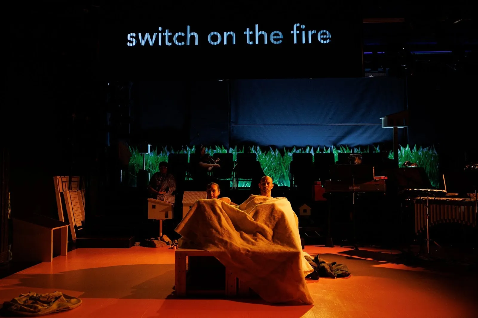 Two performers lie on a bed on stage. In the background, a musical ensemble can be seen. Projected onto the back wall of the stage is test that reads "switch on the fire."