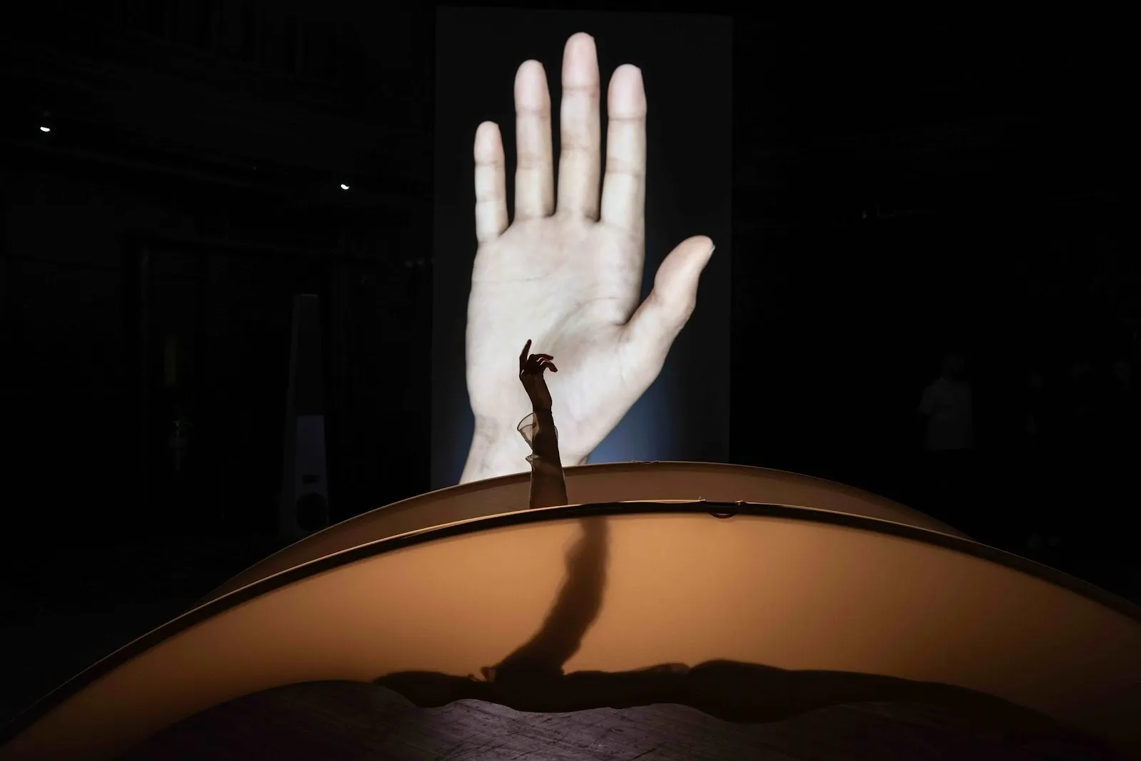 Against a large LED screen featuring the image of a raised hand, a body resting in a mesh/nylon-stretched hammock raises their hand.