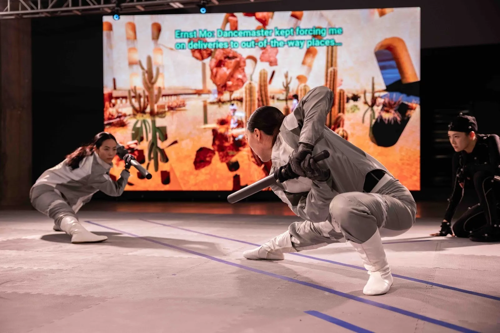 In a large space with an LED screen behind projecting an abstract world, two performers in motion-capture suits face off, crouching with one leg outstretched, pointing blunt rods at each other.