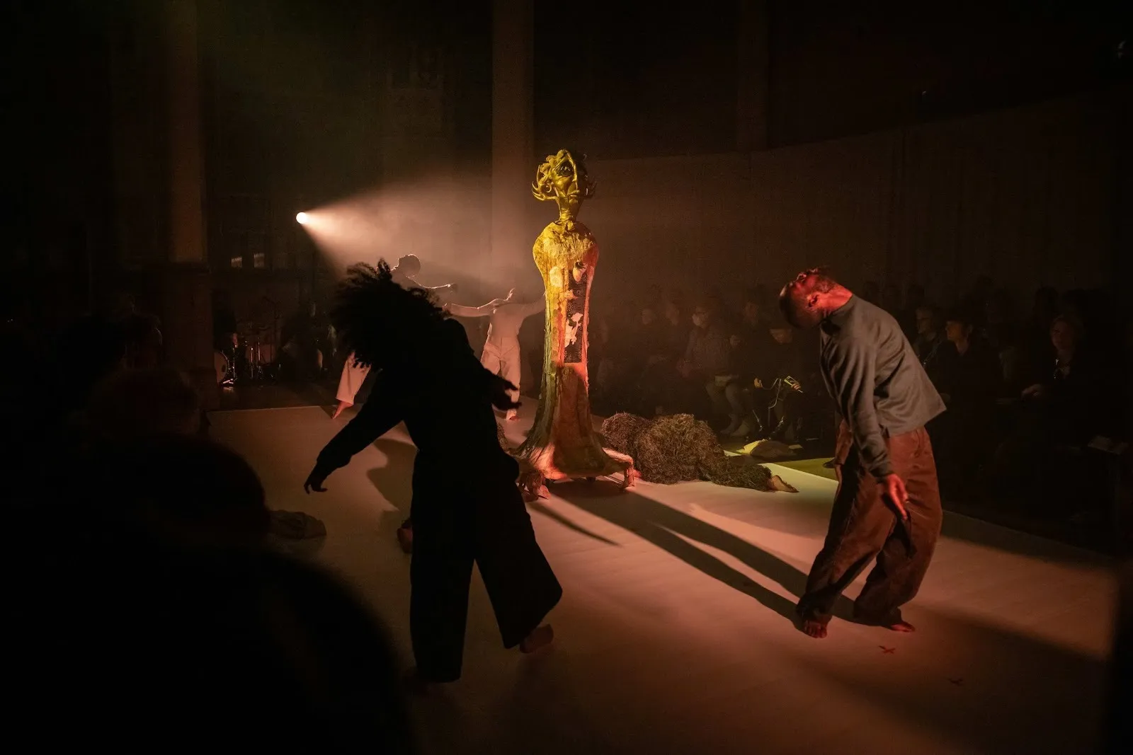 In a dimly lit space with an audience, an abstract effigy statue stands in the center, spotlit from behind. Around the statue, performers lean back in mid-movement, their heads thrown back and faces turned upwards toward the ceiling.