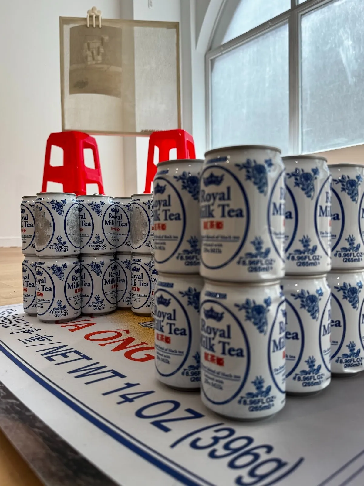 Configured stacks of Royal Milk Tea cans rest on a large printed image set on the floor of a gallery space. In the distance, another sculpture made of stacked plastic red stools supporting a transparent screen with media can be seen.