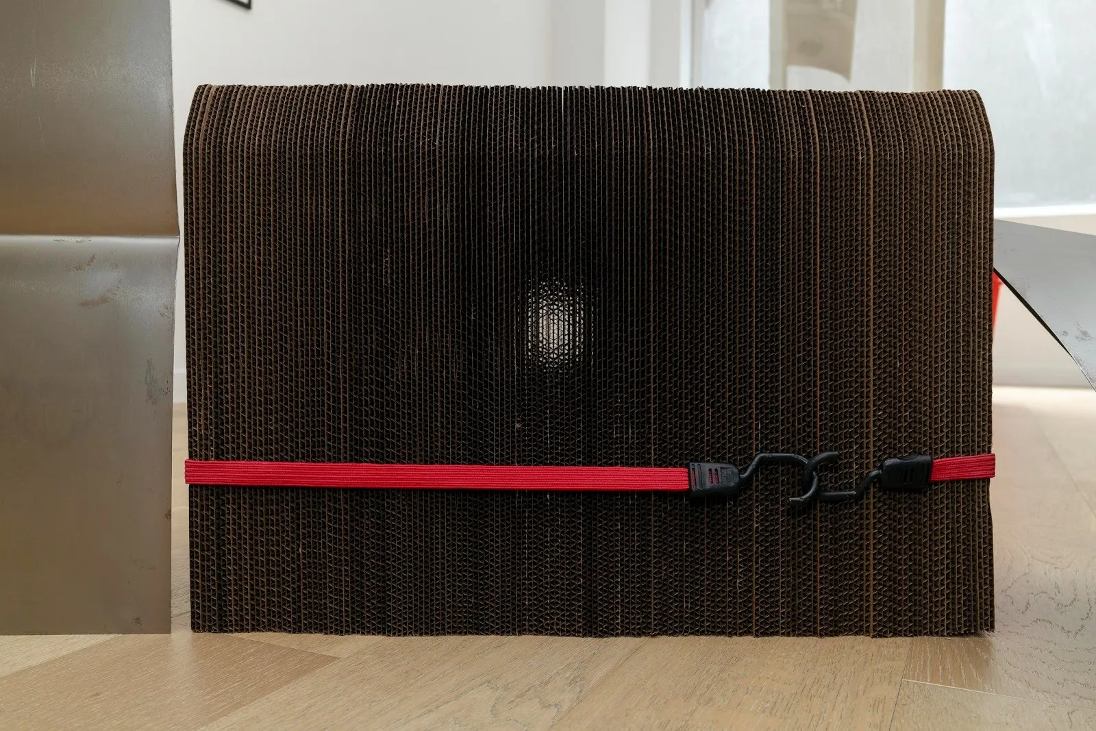 A stack of cardboard layers, all of the same size, are held together by a hooked, thick, red cord. The cardboard stack is bordered by metal sheets.