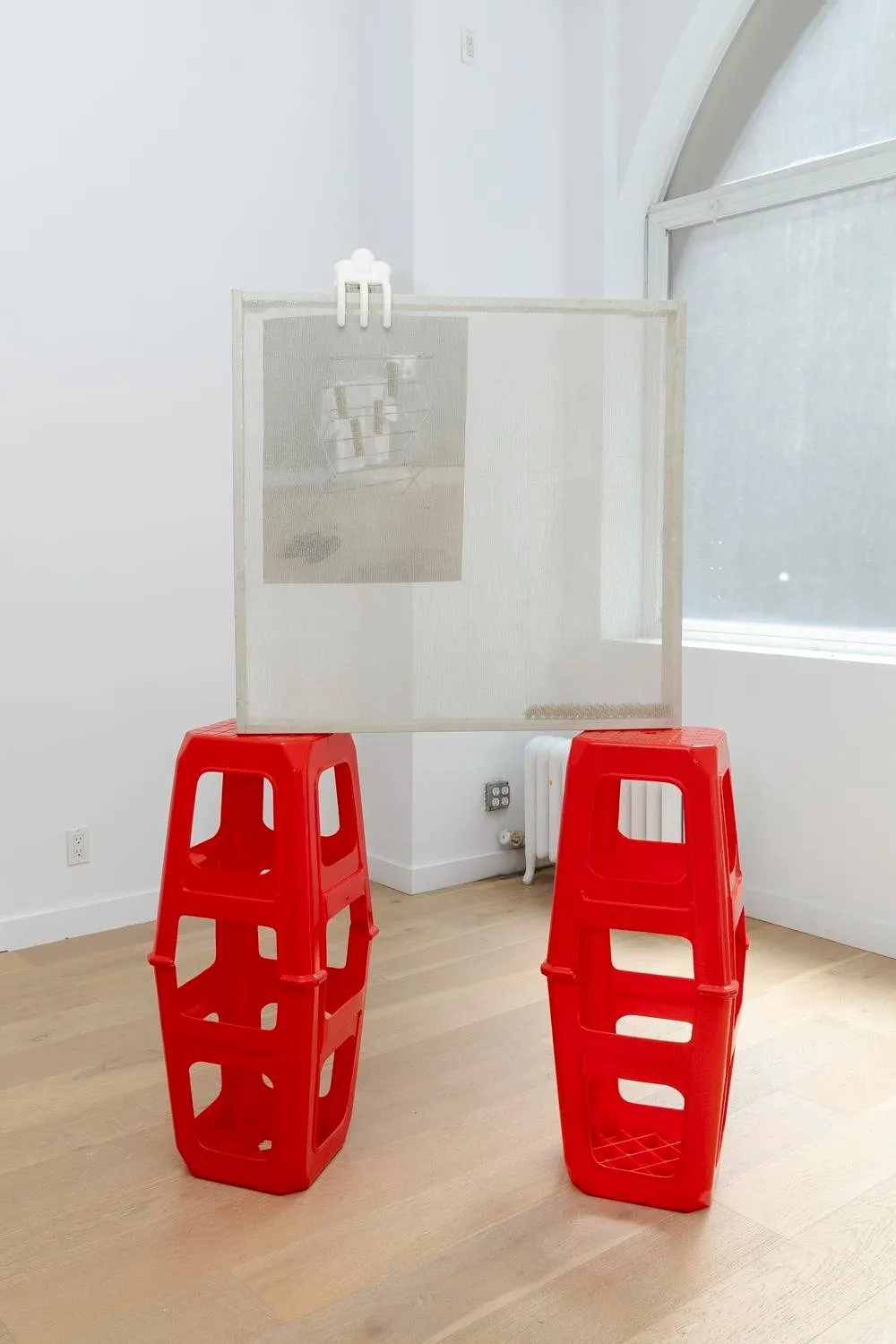 In a white gallery room, two stacks of red plastic stools support a transparent, mesh-like frame which features an image that appears to be clamped onto the frame.