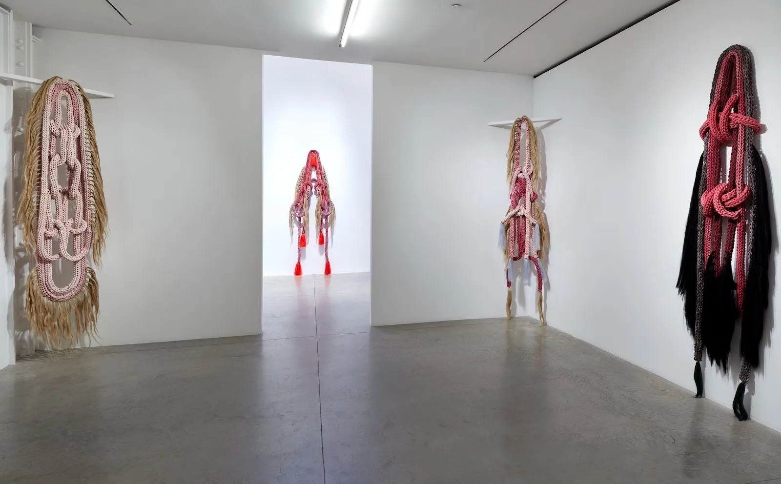 A gallery room houses sculptures made of textile and rope. Each one is of a different configuration and are all large works, and each is made of different colors.