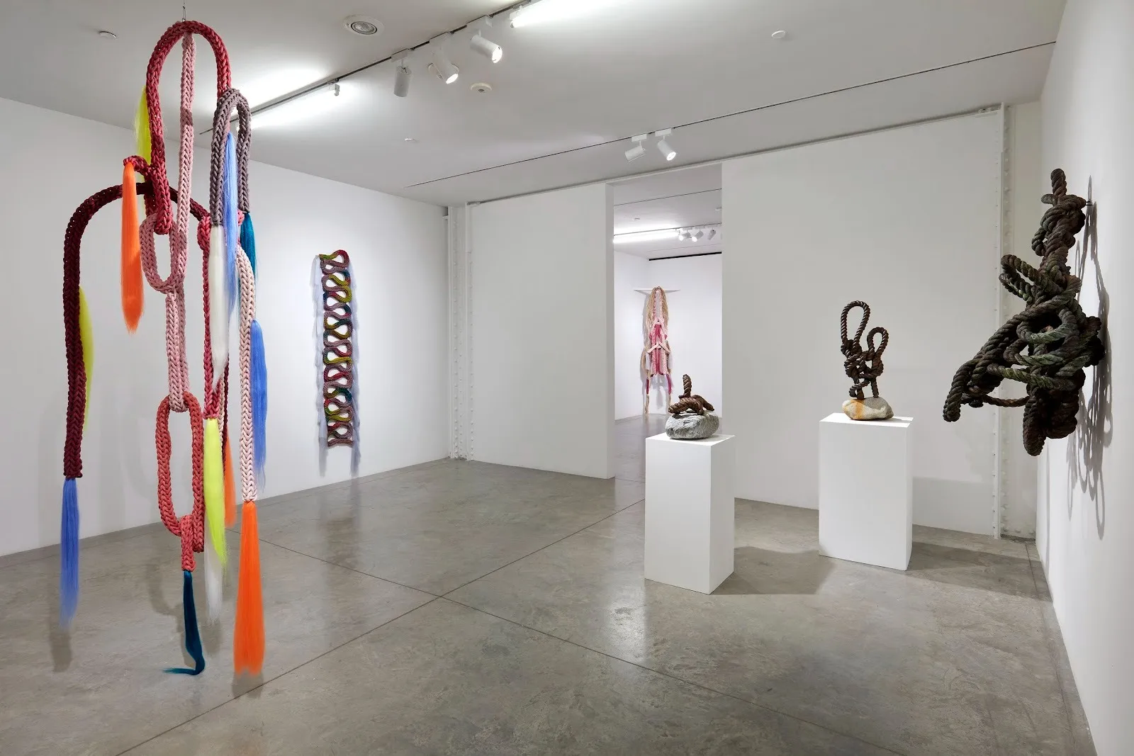 A gallery room houses sculptures made of textile and rope. Each one is of a different configuration, ranging from small to floor-to-ceiling, and each is made of different colors.