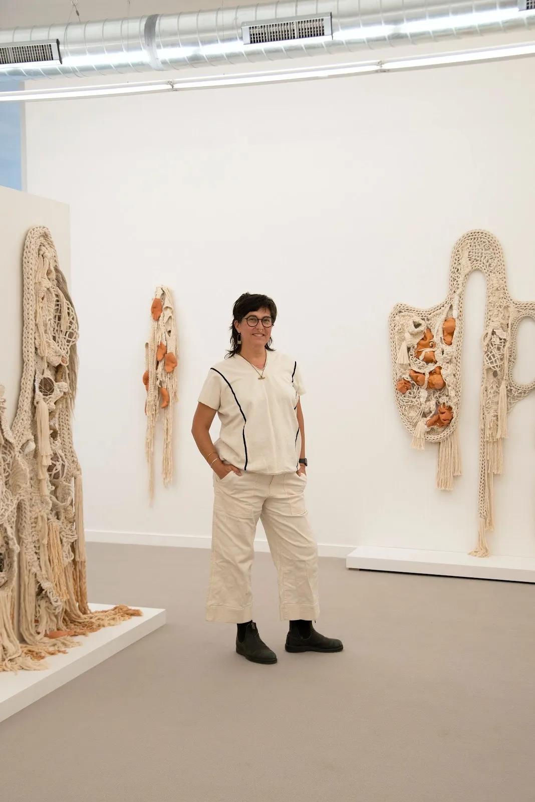 A person stands amidst rope and woven textile works mounted to the wall. Each work are of various size and shape and adhere to a beige-brown-orange color range.
