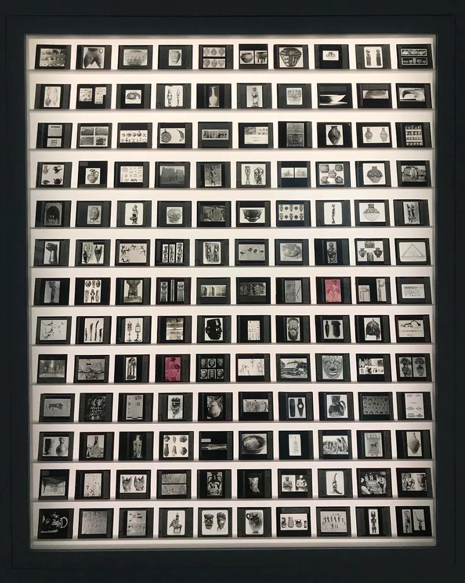 A large, backlit wall features rows of images with black paper backing. Most of the photographs are black and white, some are reddish, sepia-toned. Most of the photographs are of archeological and artistic artifacts.