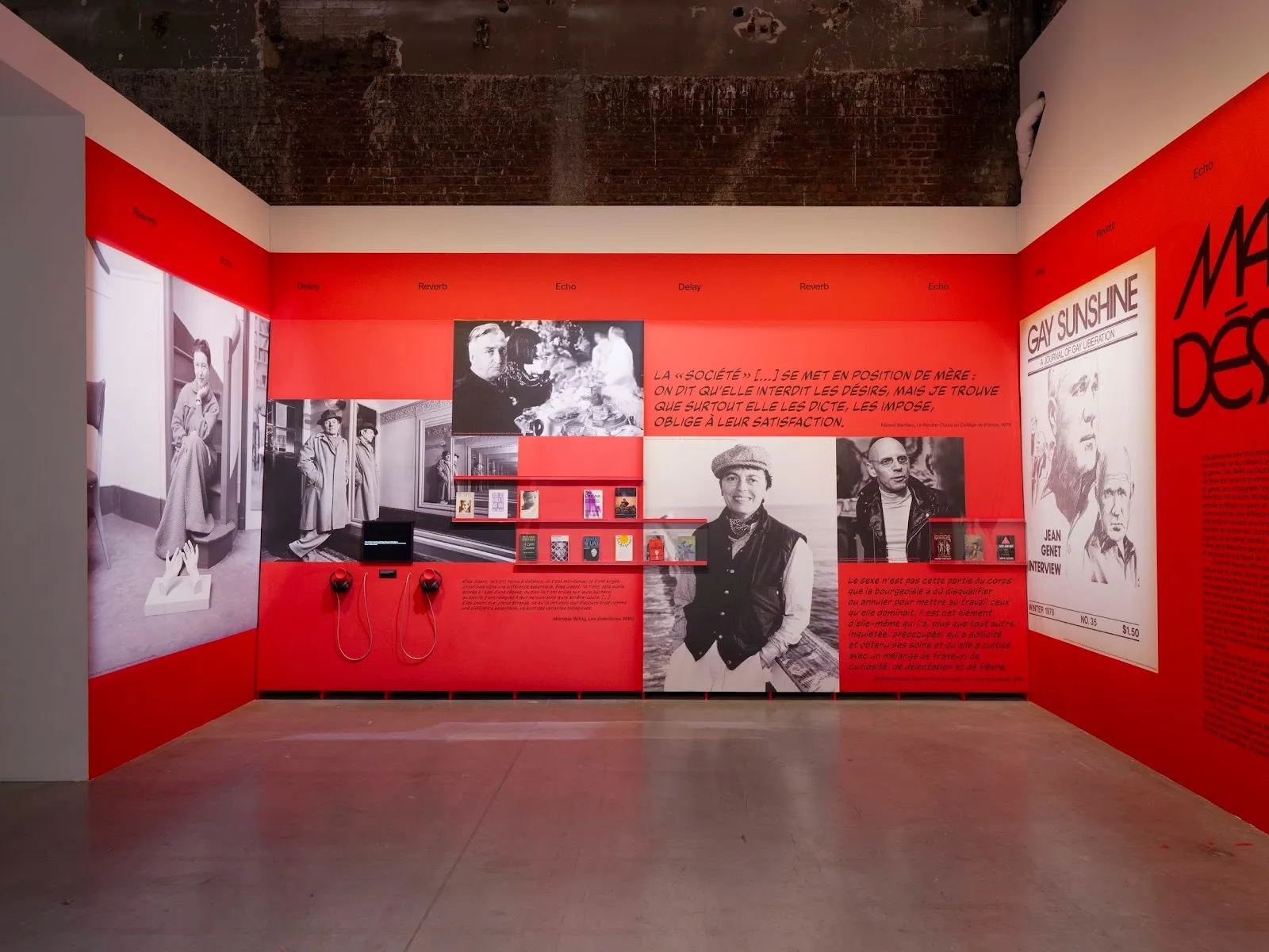 A gallery room has red walls with text vinyled on: quotes, information text. The walls also feature black-and-white photographs of various people, shelves of ephemera, and over-ear headphones.
