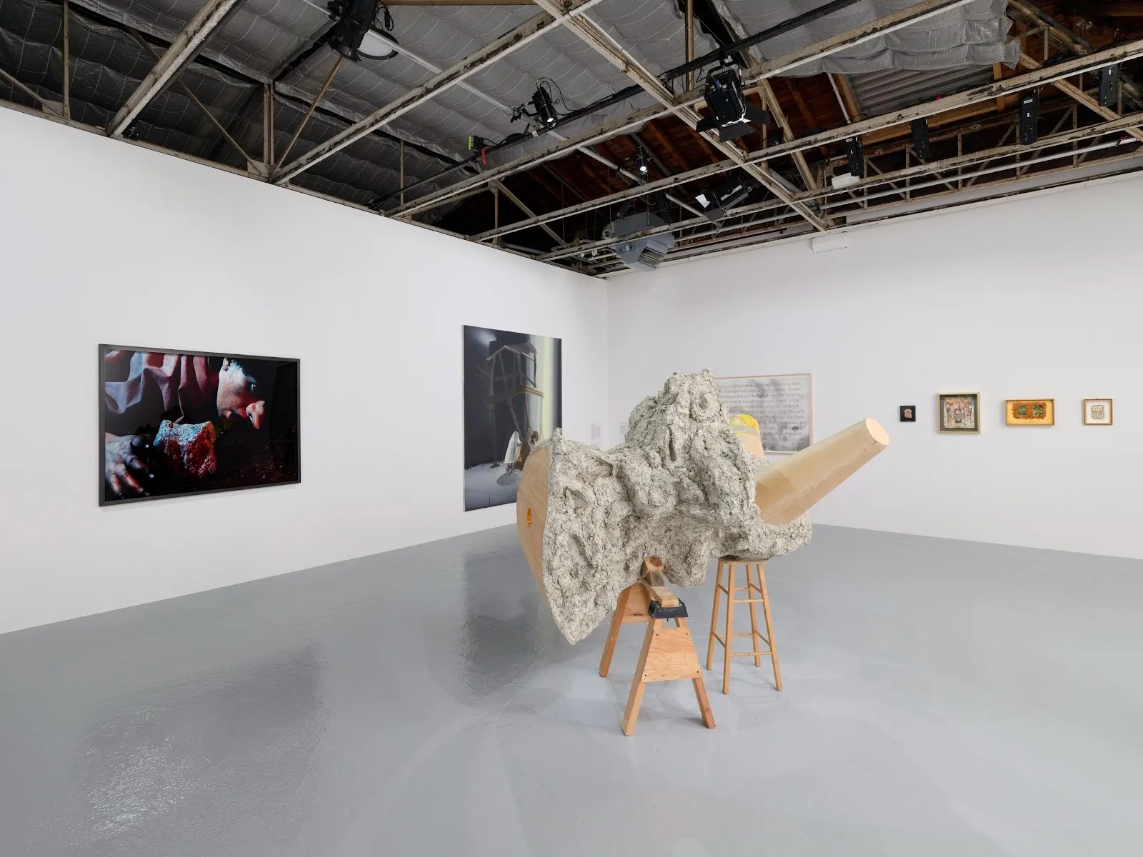 A white gallery room features a number of 2D wall works. On the floor, a sculpture made of wooden stools and a mass of lumpy, stone like material and wood sits central.