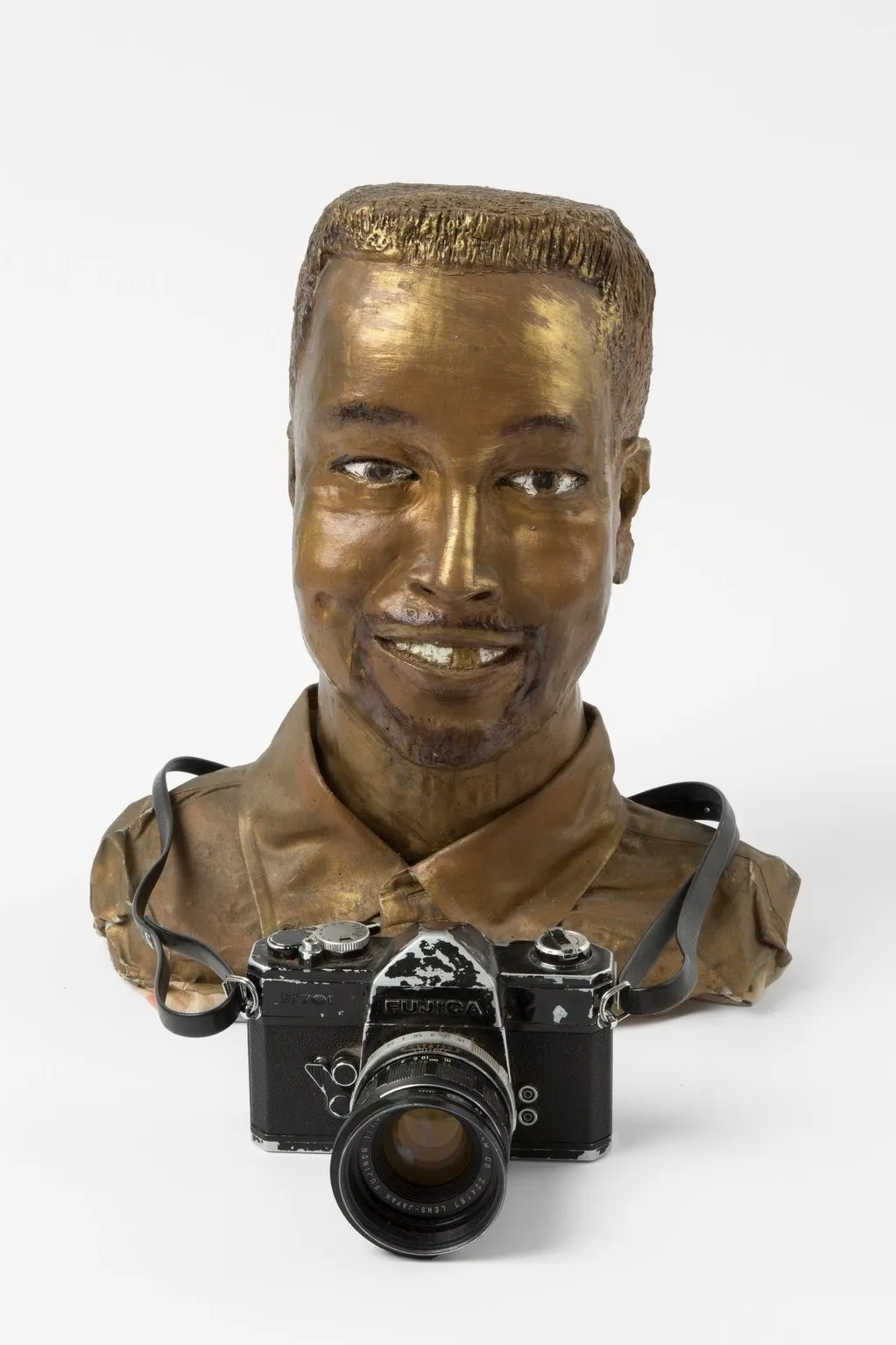 The smiling bust of a man is painted gold, in addition to an exposed front tooth. An old camera is looped around the figure's neck, standing upright in front of the bust.