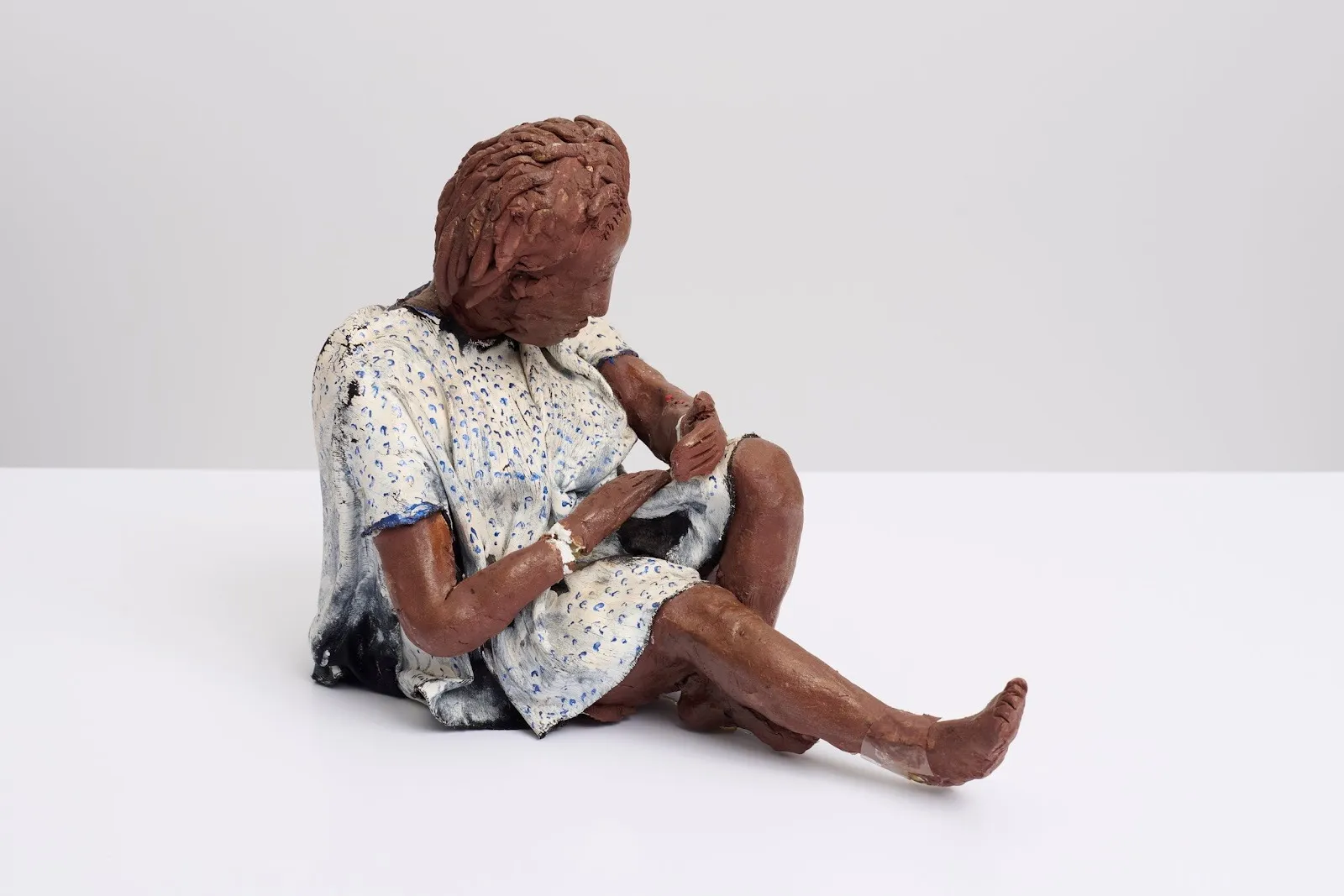 A statue depicts a figure in terra cotta finish wearing a hospital robe sitting with one knee propped up looking down at their hands.