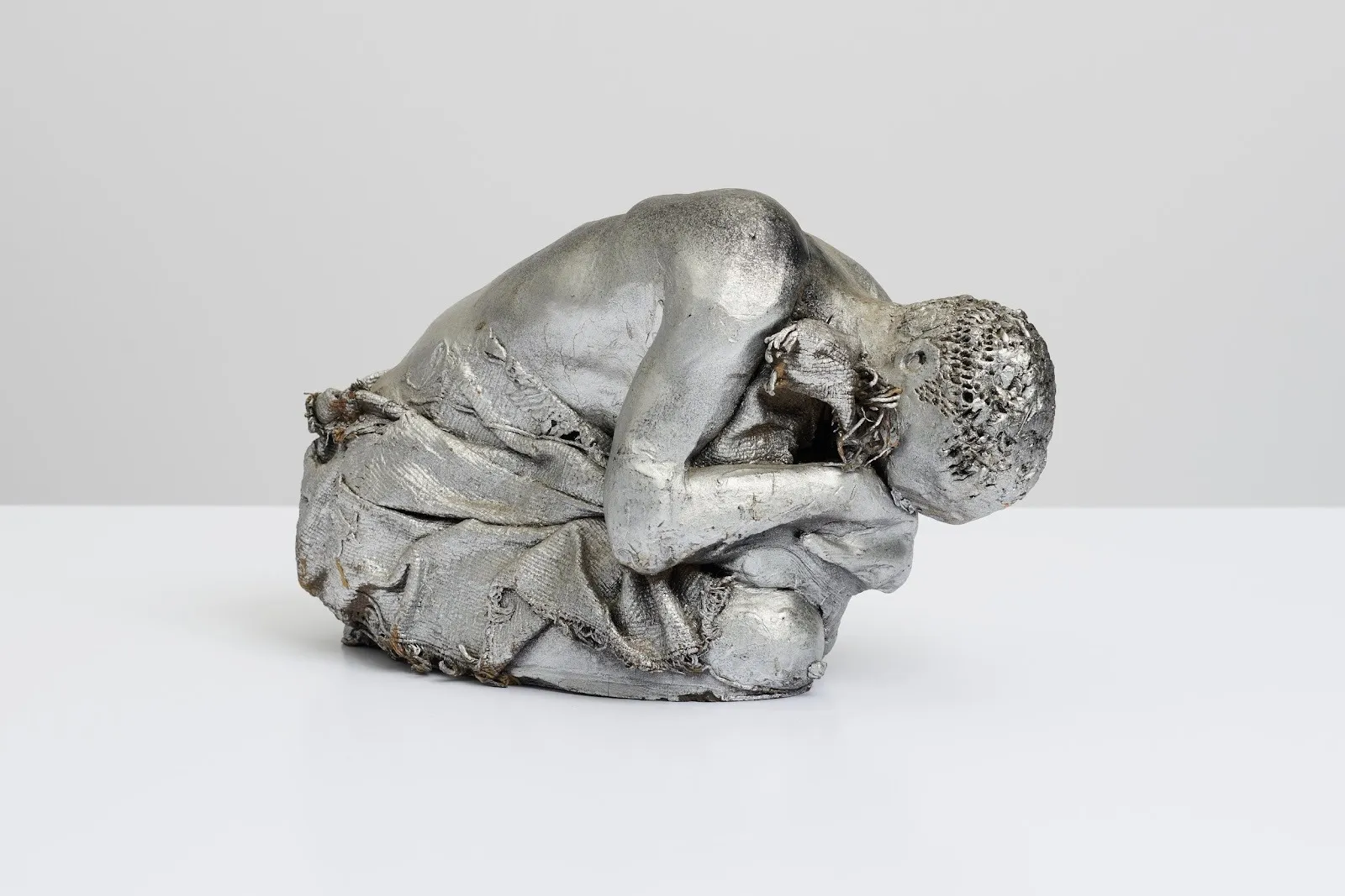 A statue features a crouched figure curled in on themselves, like an upright fetal position. Their face is obscured and torso bare. The surface of the statue is painted with silver pigment.