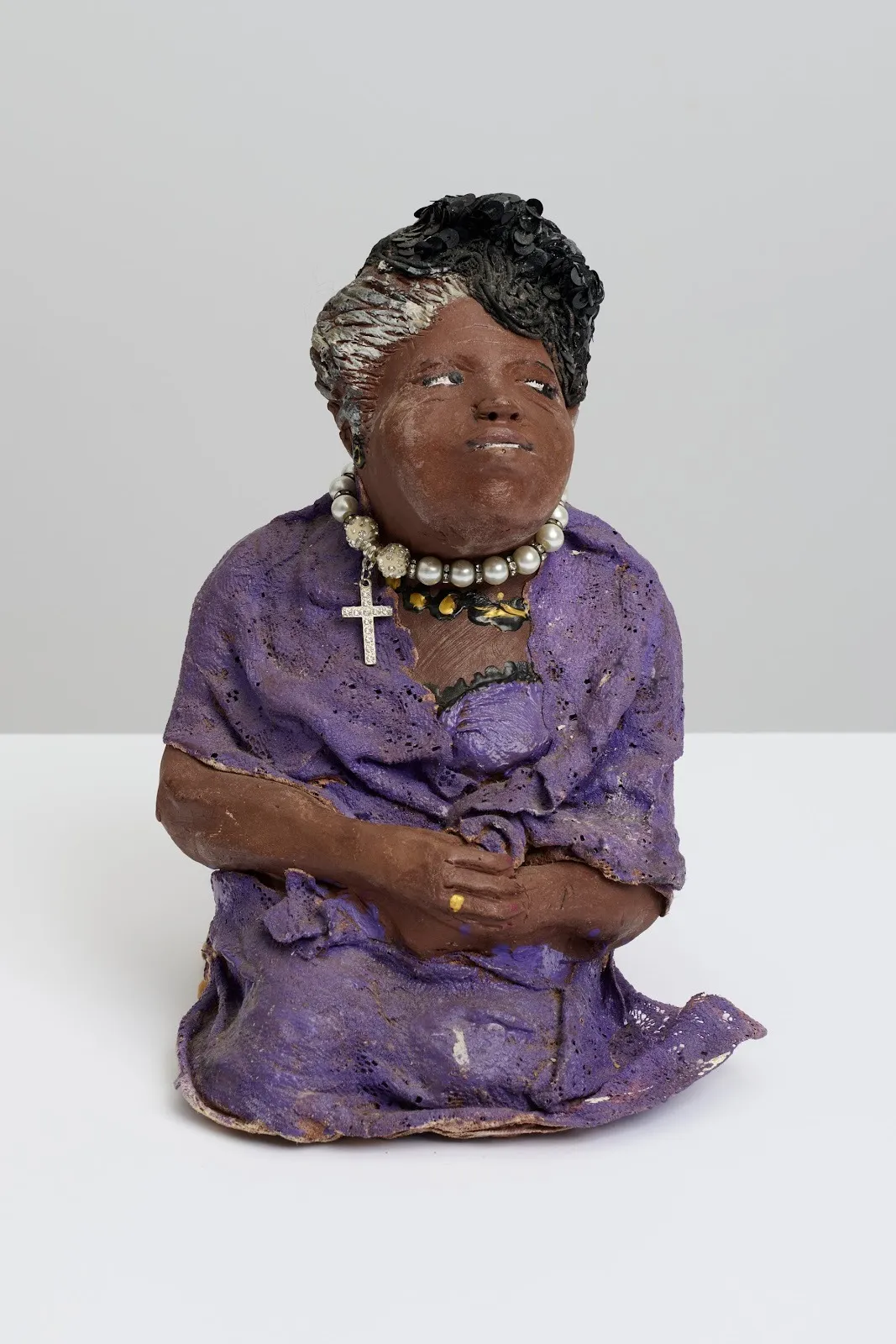 A votive-like statue features a woman in a purple dress kneeling with her hands clasped together. She smiles slightly and looks to the side. She wears a large pearl necklace with a cross charm.