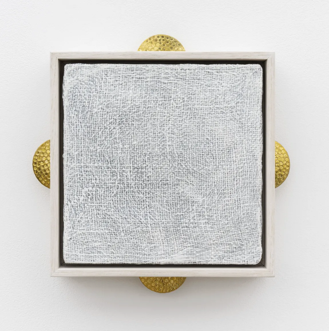 A square canvas painted white sits within a thin, white wooden frame mounted to the wall. Behind the frame, a semicircle of hammered.dotted bronze semicircles peek out, one on each side.