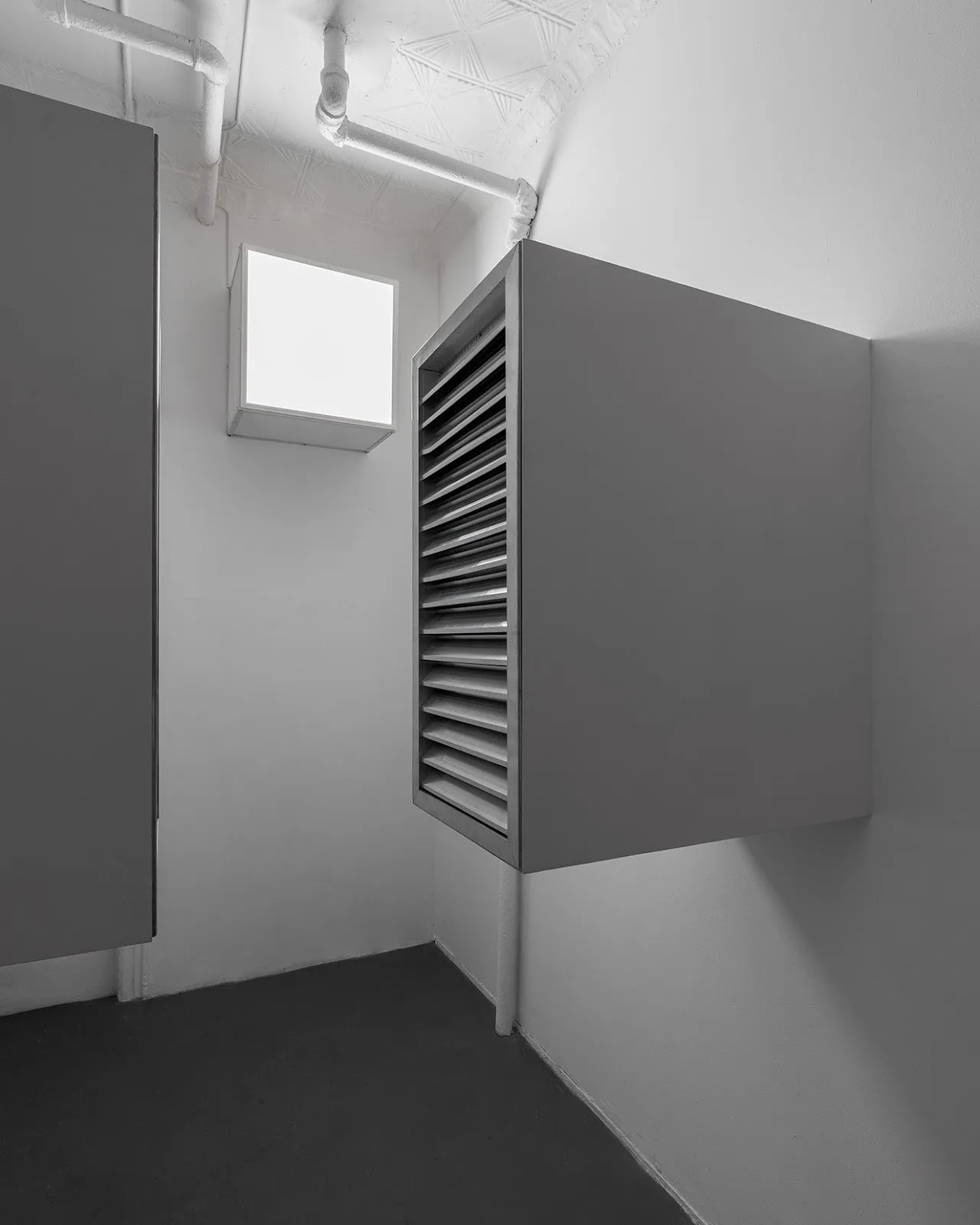Close-up of a single wall-mounted louvered box in a minimal white room with exposed pipes.