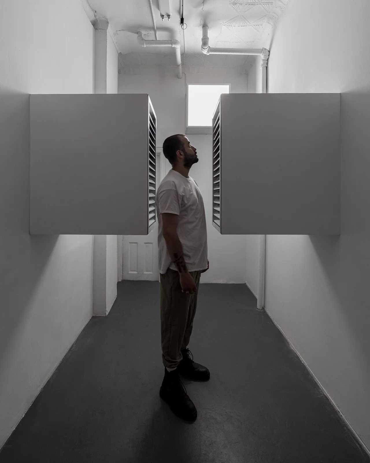 Person standing between two large wall-mounted boxes in a narrow white room, looking up.