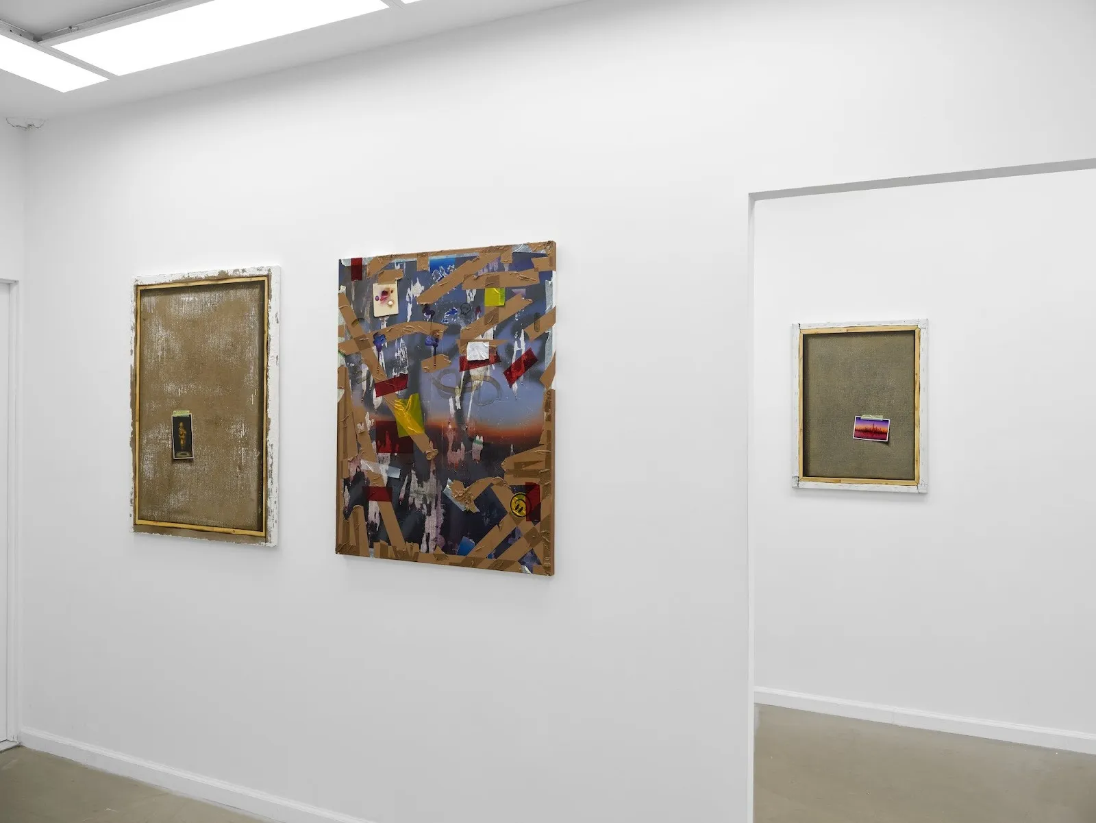 Gallery wall displaying three mixed-media works with textured surfaces, collage elements, and postcards or stickers affixed to their centers.