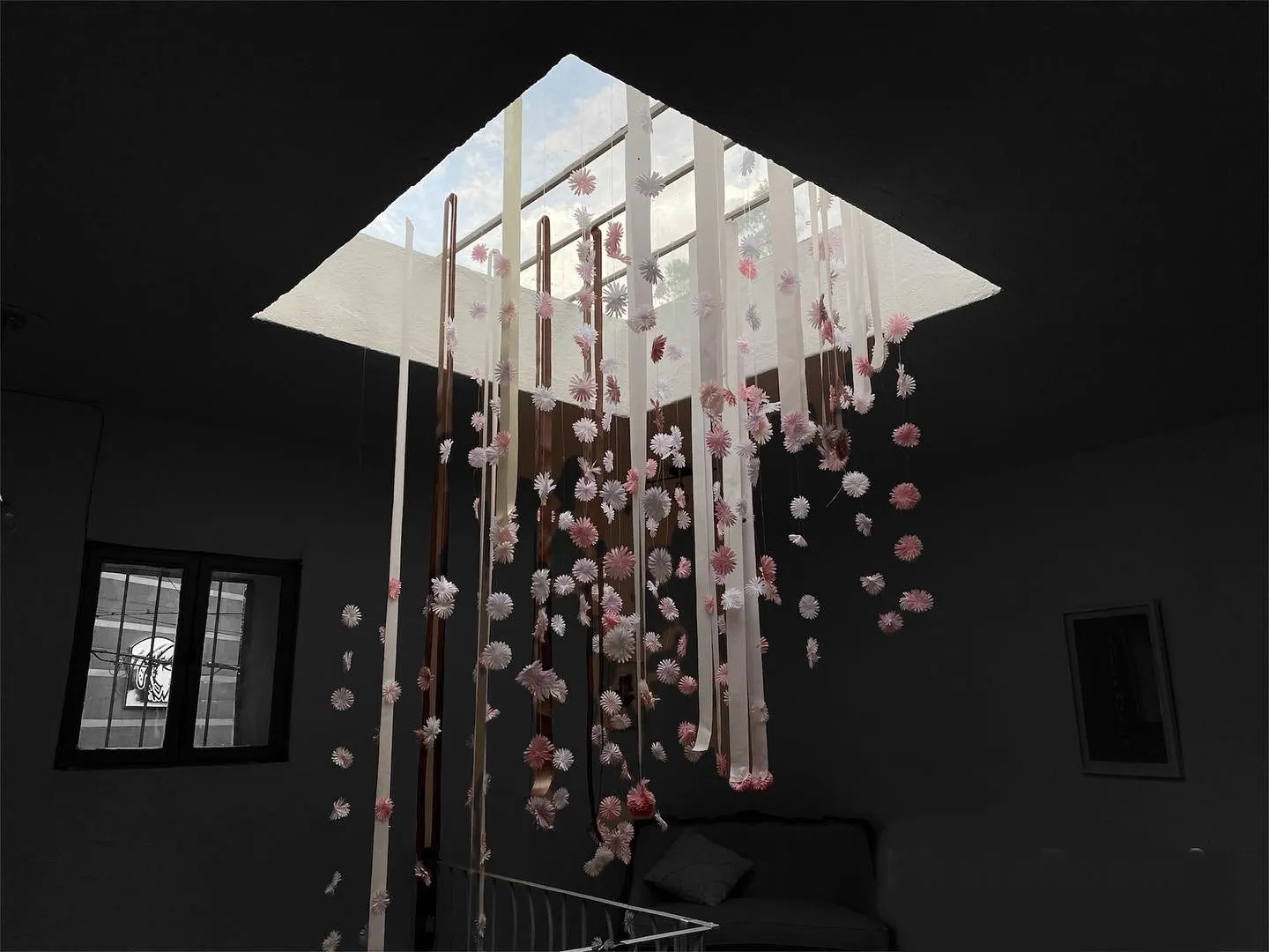 Hanging installation of delicate pink and white flowers suspended from a skylight, cascading into a dimly lit room.