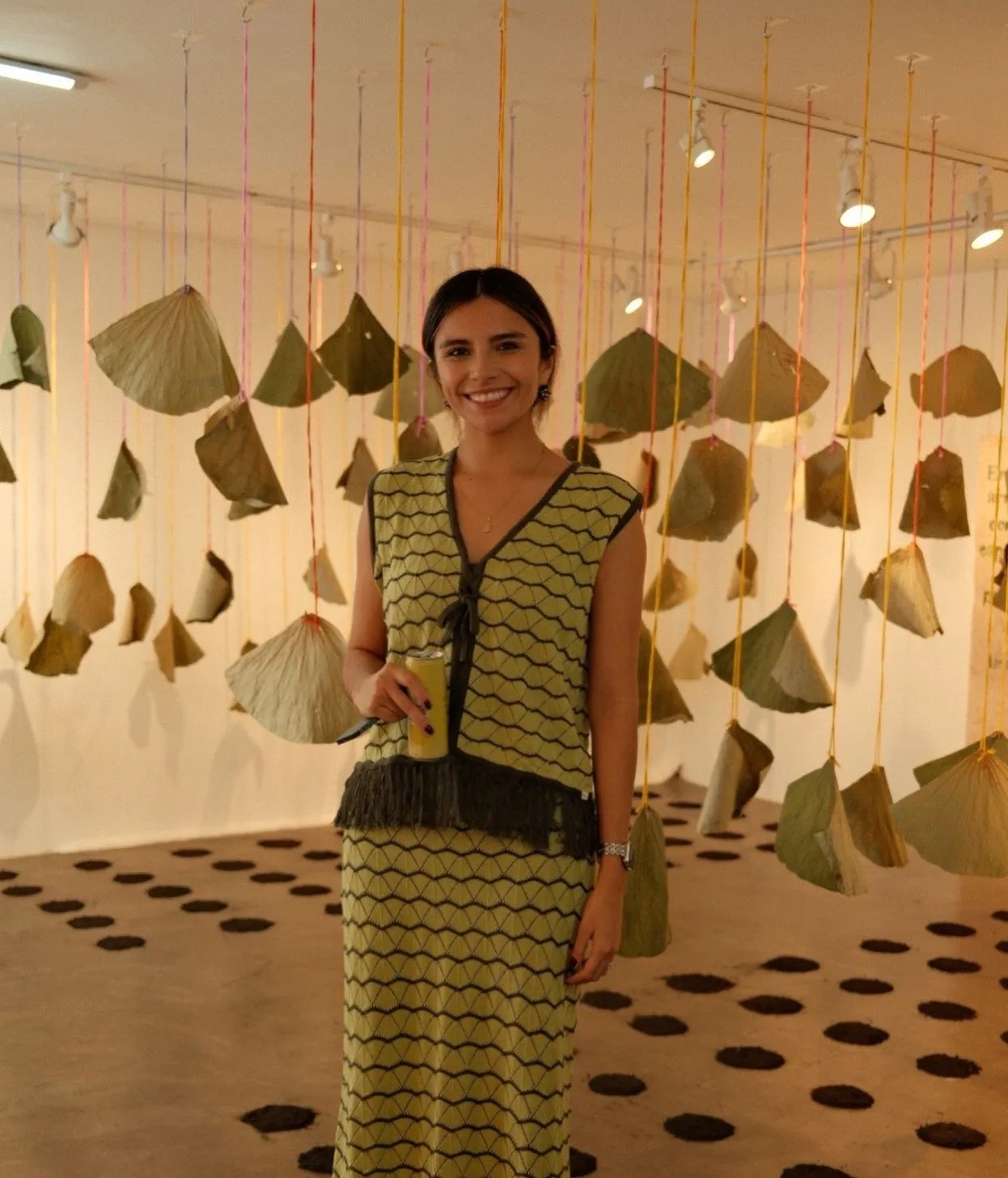A smiling woman stands in a gallery with hanging dried leaves and colorful strings, wearing a matching patterned outfit and holding a drink.