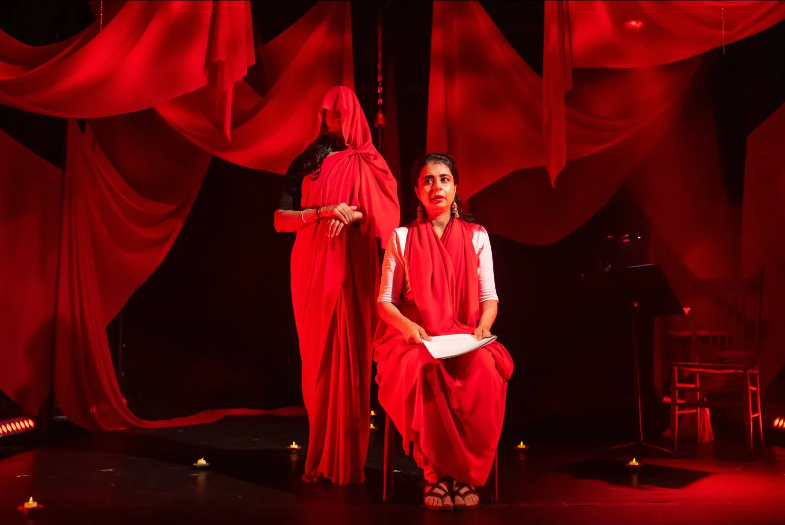 A dramatic theatrical scene bathed in red light, featuring two women in red draped garments—one seated and holding a script, the other standing behind with her face covered—set against flowing red fabric and small candle lights.