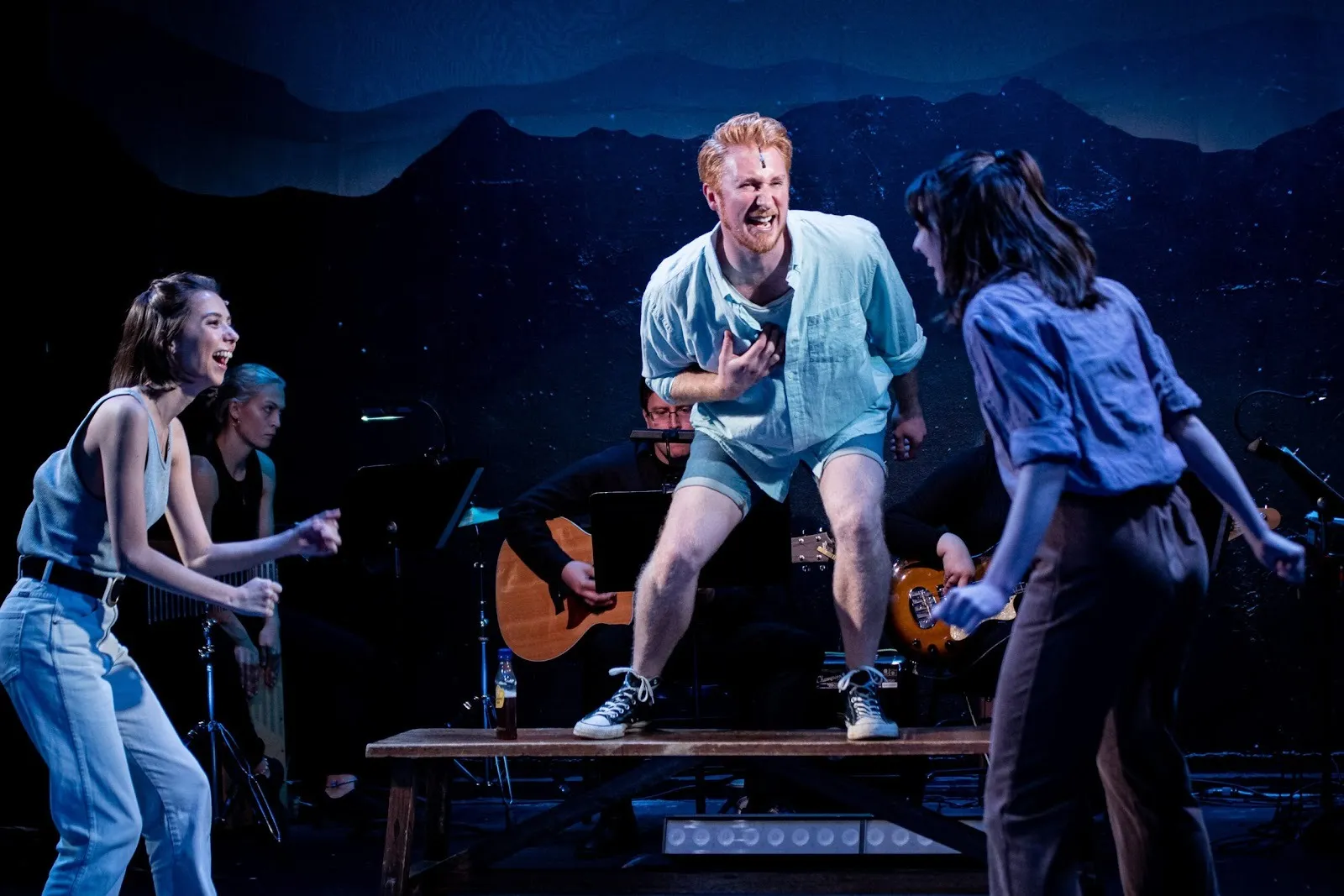 A lively musical performance with three actors in energetic motion; one man stands on a bench passionately singing while musicians play in the background against a mountain-themed backdrop.