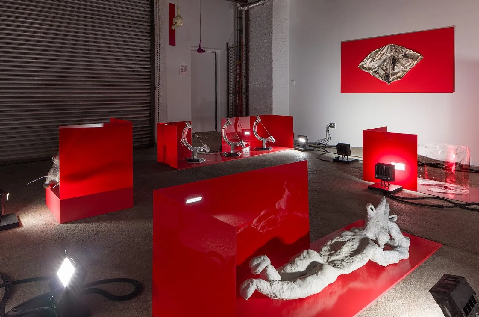 Installation view of an art exhibition featuring glossy red display structures, metallic sculptures, and dramatic lighting in an industrial space.