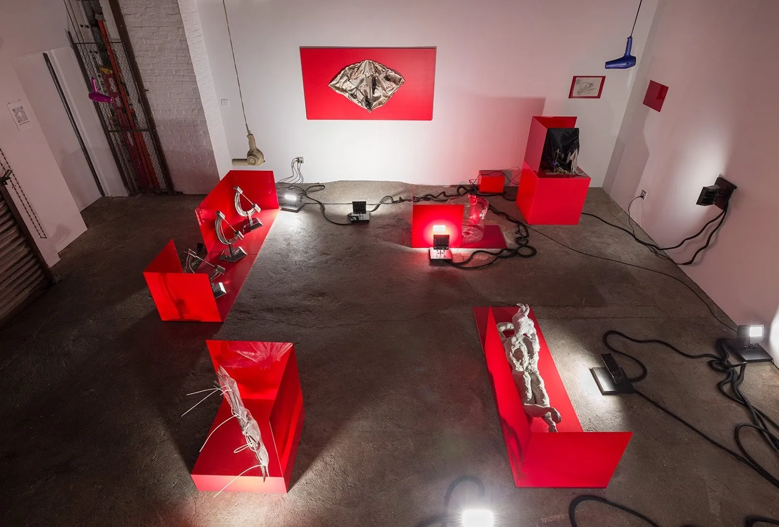 Installation view of an art exhibition featuring glossy red display structures, metallic sculptures, and dramatic lighting in an industrial space.