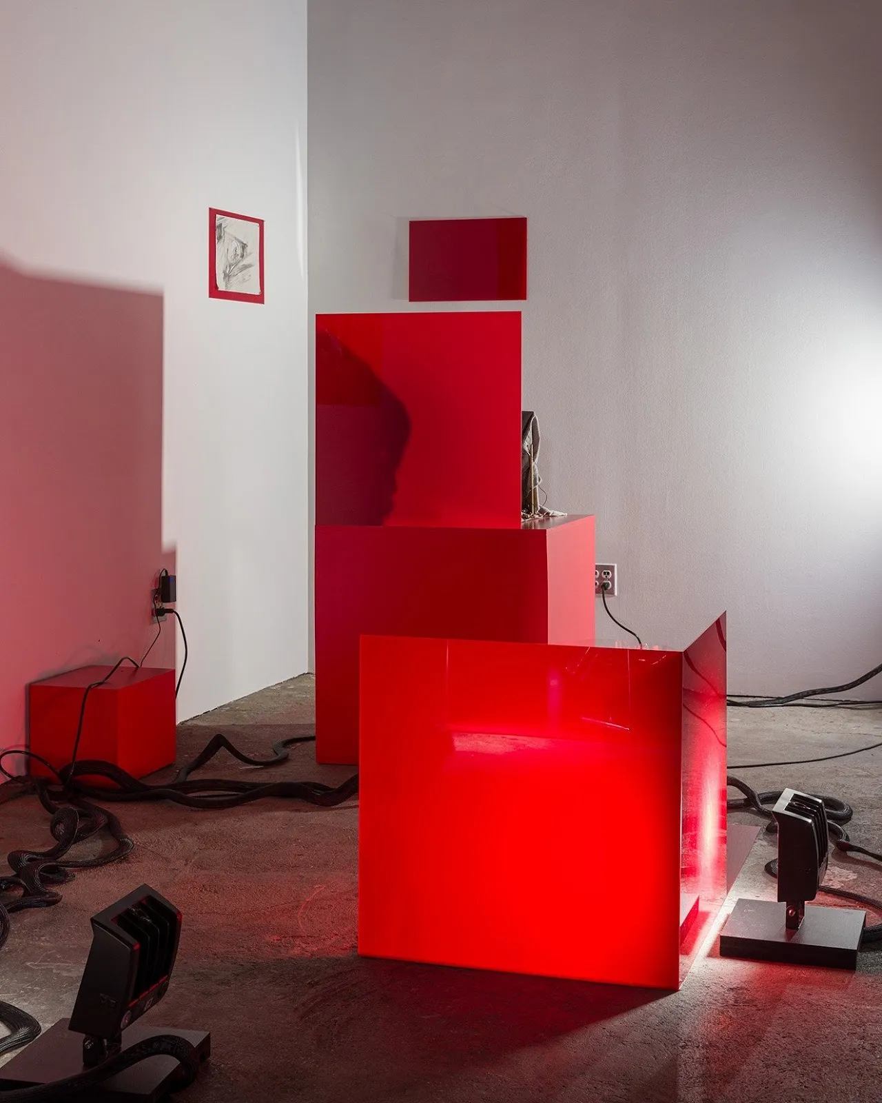 Installation view of an art exhibition featuring glossy red display structures, metallic sculptures, and dramatic lighting in an industrial space.